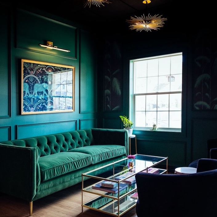 A dramatic nighttime view of the Rhino Wine Bar’s emerald green lounge features a central framed artwork of two elephants under a sunburst motif, backlit by a golden picture light. The green velvet sofa below mirrors the wall tone, creating a seamless visual effect. Brass starburst chandeliers overhead add an extra layer of drama and glamor. This interior showcases the boutique design studio’s creative expertise, curating layered textures and rich hues to craft a distinctive atmosphere for wine lovers in the Twin Cities.