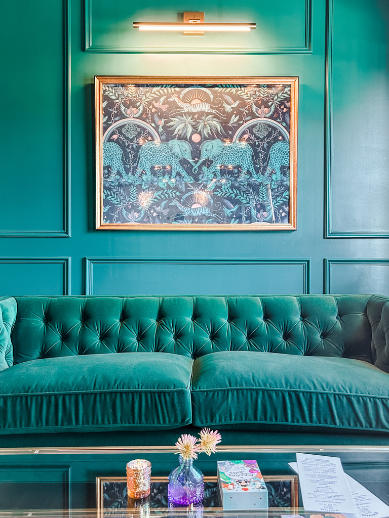 A framed artwork featuring twin elephants with ornate detailing is centered above a deep emerald green velvet sofa. The surrounding paneled wall is painted the same rich emerald tone, while a picture light above the frame casts a warm glow on the eclectic scene of jungle flora and fauna. A glass coffee table in the foreground displays small votive candles and purple blooms in a mini bud vase. Located in the Minneapolis-St. Paul area, this cozy lounge corner balances bold color with whimsical decor for an unforgettable wine bar experience.