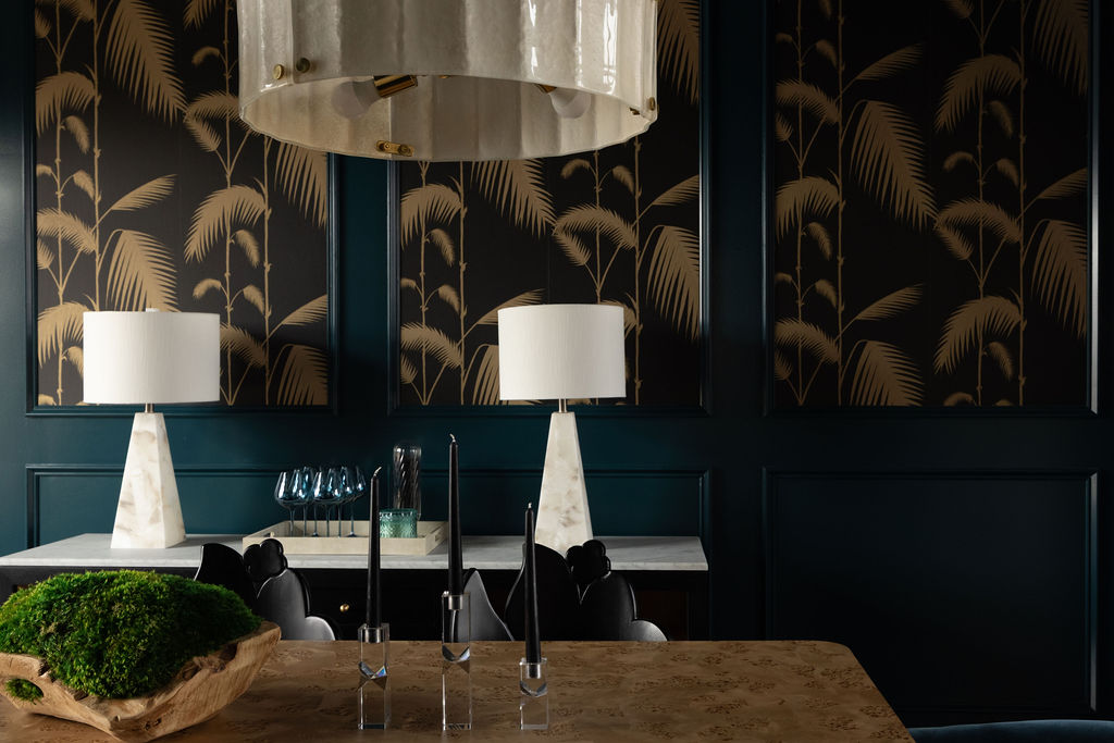 Detail view of a moody dining room featuring dark teal paneled walls, botanical wallpaper panels, a stone dining table, sculptural black chairs, and modern lamps.