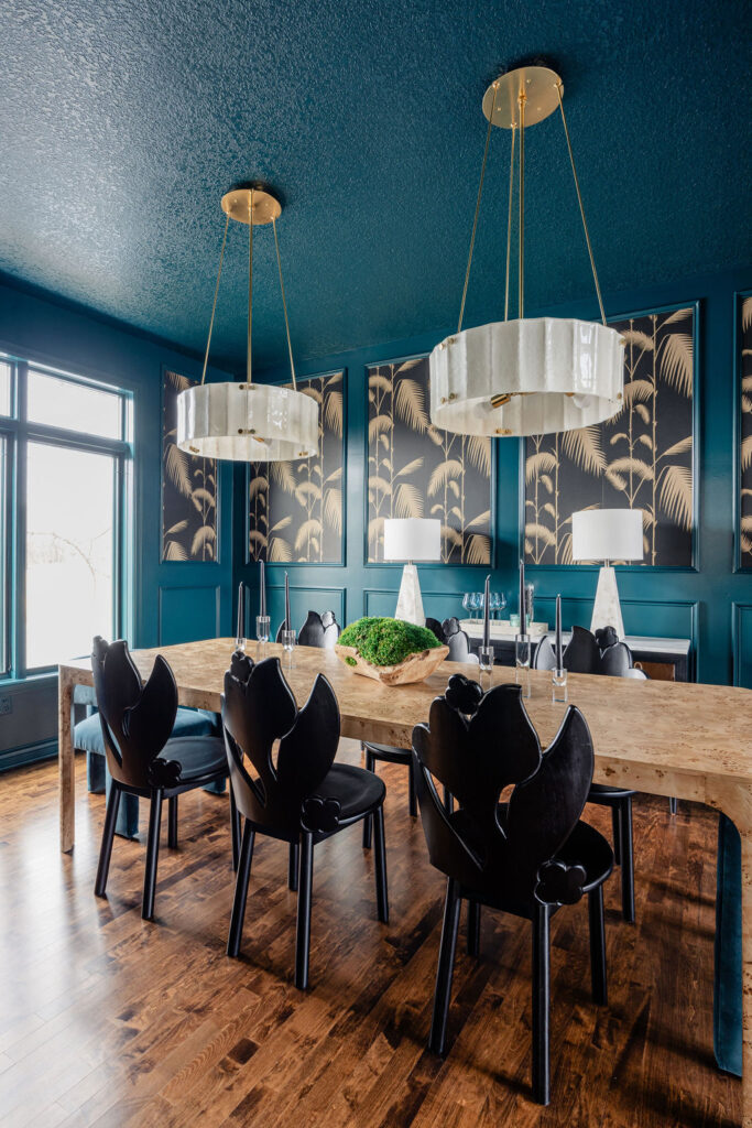 Moody dining room featuring teal paneled walls, framed botanical wallpaper, a stone dining table, sculptural black chairs, and modern pendant lights.