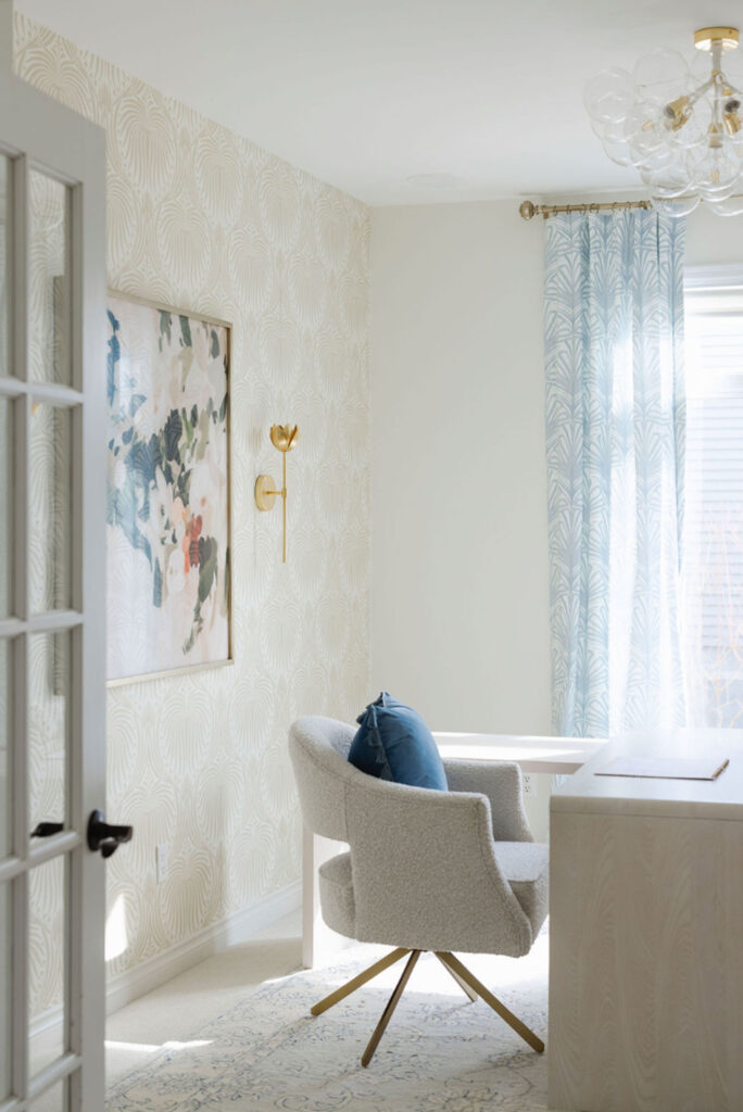 Light-filled home office featuring a white desk, textured upholstered chair, soft patterned wallpaper, brass wall sconce, and sheer blue curtains.