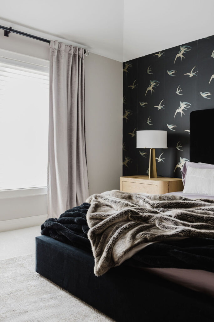 A modern bedroom with a black upholstered bed layered in plush blankets, a light wood nightstand with a brass lamp, soft gray curtains by a large window, and dark wallpaper patterned with flying birds.