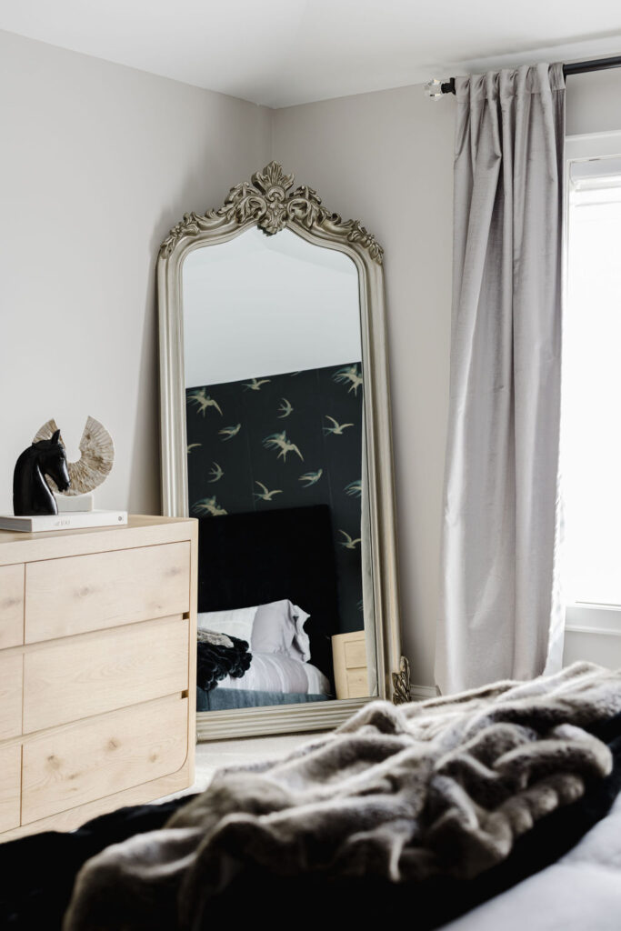 An ornate full-length mirror leaning beside a light wood dresser reflects a bed with dark bird-patterned wallpaper, while gray curtains frame a sunlit bedroom window.