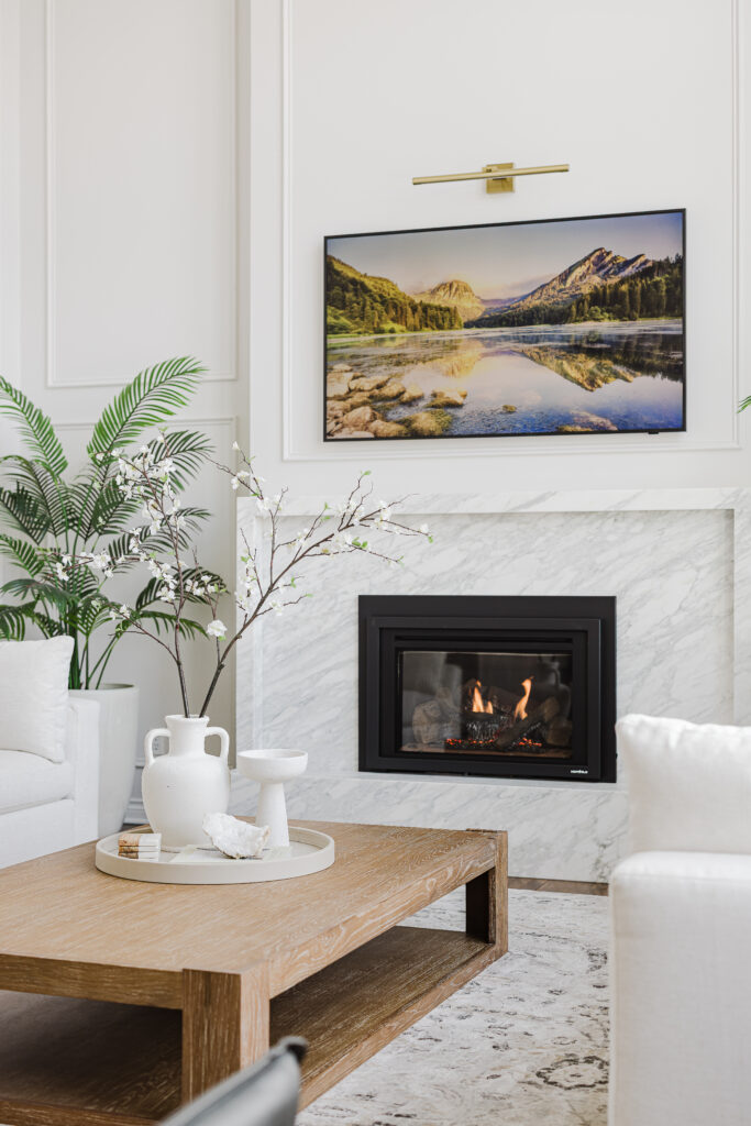 Living room remodel featuring a white marble fireplace, wall-mounted TV, brass picture light, and modern neutral decor.