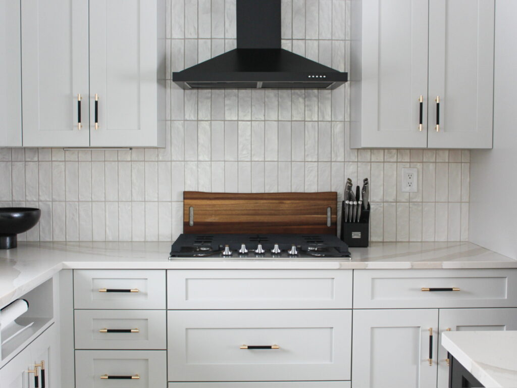A close-up of a modern kitchen cooktop area in a Minneapolis-St. Paul home, featuring soft gray shaker cabinets with black and gold hardware. The matte black range hood is centered above a five-burner gas cooktop, framed by shimmering vertical tile backsplash and a warm walnut knife block and cutting board. The mix of sleek finishes and rich textures creates a grounded yet elevated cooking space. Firelight Design’s thoughtful layout and styling reflect their boutique approach to modern interiors, balancing warmth, function, and visual simplicity.