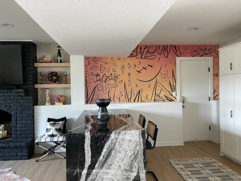 A bold lower-level bar area in a Minneapolis-St. Paul home features a dramatic black marble waterfall island with tan veining, paired with cane-backed barstools. The wall behind the bar showcases a custom orange-to-pink gradient mural with expressive line art and graffiti-style symbols. Built-in shelves hold sculptural pottery and colorful books, while a matte black painted brick fireplace adds contrast. The Firelight Design team brought their creative vision to life with curated decor and one-of-a-kind finishes, blending Southwest flair with contemporary artistry.