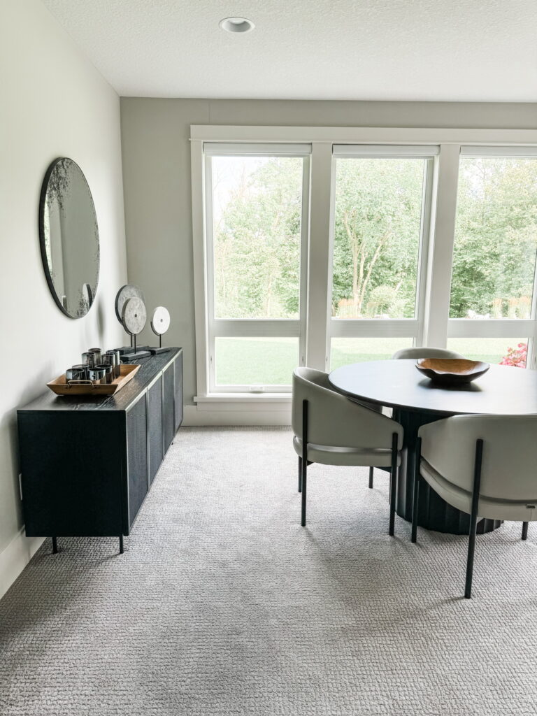 A serene dining nook in a Twin Cities home displays a round black dining table surrounded by minimalist taupe leather chairs on a soft neutral carpet. A sleek black sideboard topped with sculptural decor sits beneath a round mirror, with floor-to-ceiling windows framing a view of lush greenery. The boutique design firm’s expertise shines in the clean, open layout—ideal for accessibility and visual harmony.