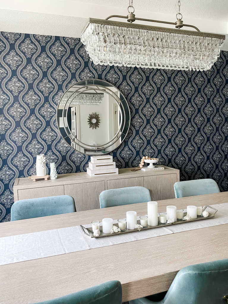A full dining room view highlights a long, light wood table with a white runner and a centerpiece of white candles in assorted sizes. The surrounding pale blue velvet chairs contrast beautifully against a navy damask-patterned wallpaper. Above the sideboard, a round silver mirror and a rectangular crystal chandelier add elegance and reflectivity. The look balances traditional decor with clean modern lines.