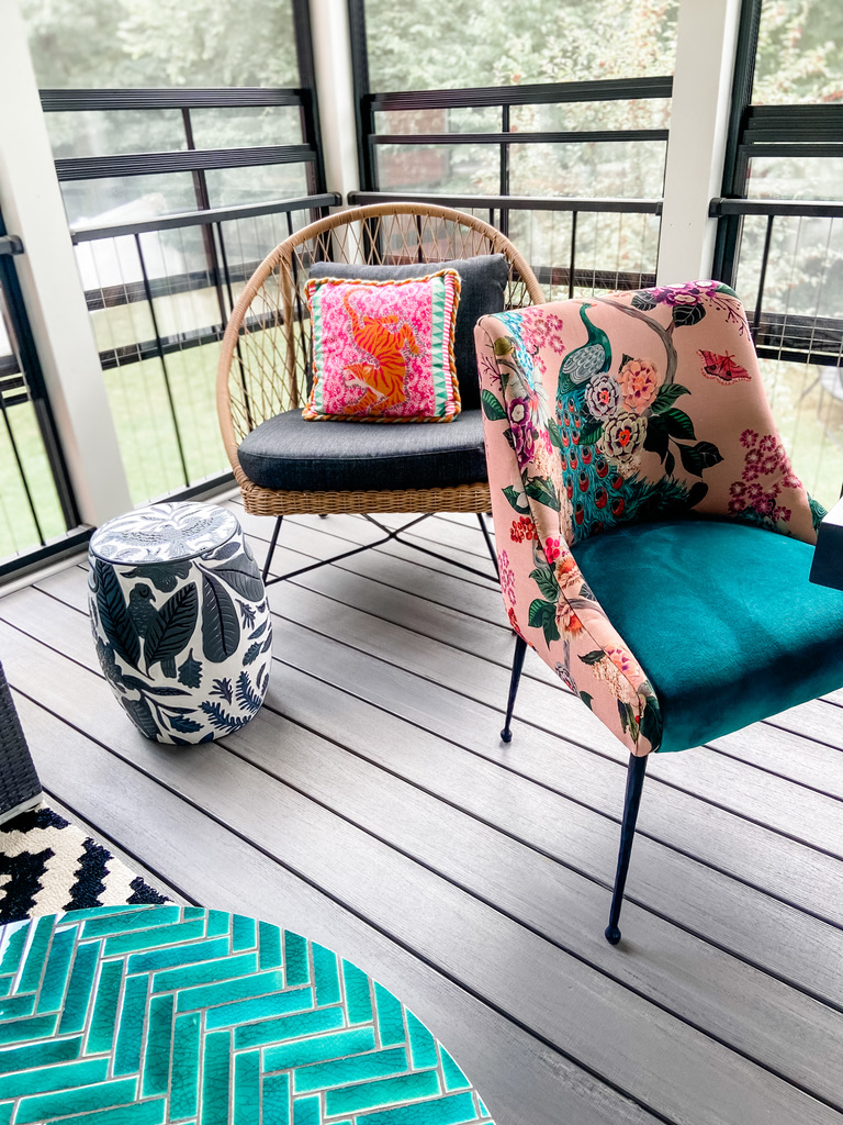 A close-up of a vibrant screen porch in the Minneapolis-St. Paul area shows a round wicker chair with a charcoal cushion and a bright coral pink pillow, paired with a pink floral upholstered chair featuring a peacock motif and teal seat. A blue and white ceramic side table and emerald green herringbone tile coffee table add rich texture. This lively, colorful space blends comfort and playful design with accessible walkways.