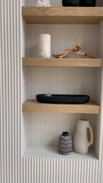 This close-up of a built-in shelving nook features three floating wood shelves set against vertical white fluted paneling. The shelves are styled with sculptural vases, driftwood, and minimalist black and white ceramic pieces. The neutral tones and varied textures add a modern, curated feel to this compact living room detail.
