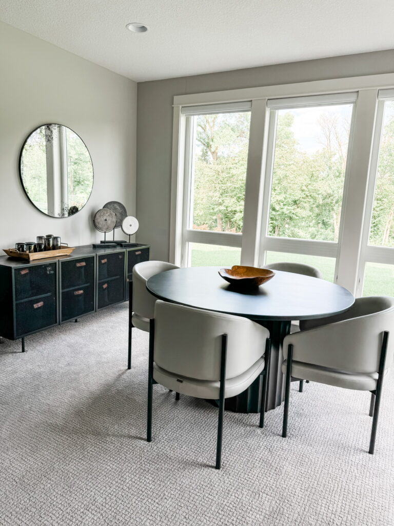 A sophisticated corner of the living room features a round black pedestal table surrounded by four ivory barrel chairs. Natural light pours in through floor-to-ceiling windows with a lush backyard view. A black sideboard with leather-pull drawers sits beneath a large circular mirror, with modern sculptures and bar accessories placed on top. This serene space exemplifies the Minneapolis interior design team’s commitment to combining functionality and beauty with modern silhouettes and natural light.