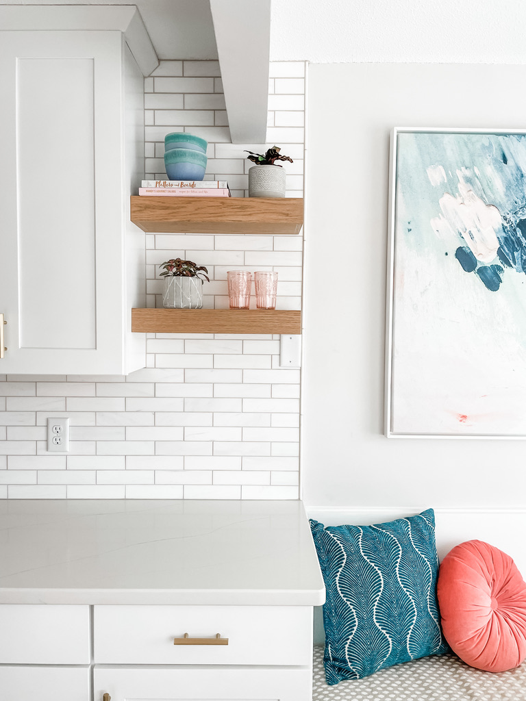 A bright and airy kitchen corner features a white quartz countertop with sleek white cabinetry and brushed gold hardware. Above the counter, two floating wooden shelves display small ceramic planters, decorative books, and pink glass tumblers. The white subway tile backsplash extends to the ceiling, creating a clean backdrop. On the adjacent bench seat, a teal throw pillow with a wave pattern and a round coral pillow add pops of color. A large abstract painting in teal, white, and blush hues hangs nearby.