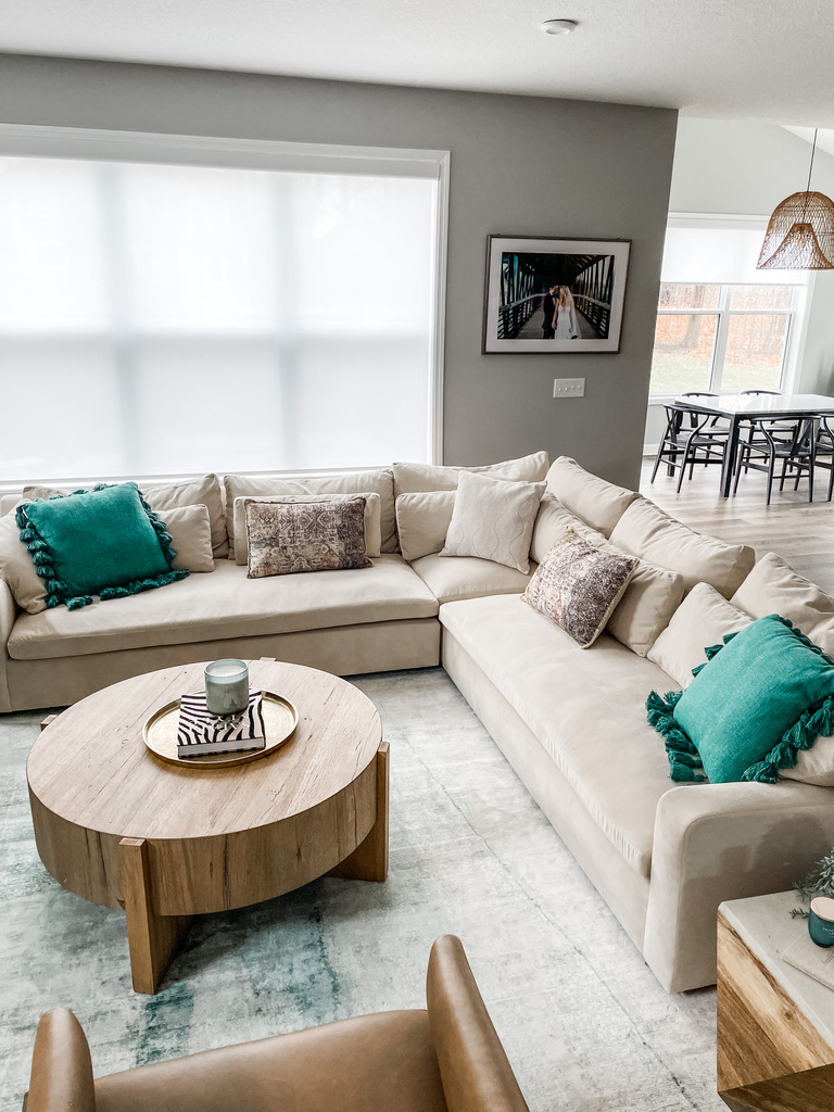 A bright, welcoming living room centers on a large L-shaped cream sectional layered with textured pillows in emerald green and patterned neutrals. A round wood coffee table holds a gold tray, zebra-striped book, and a glass candle. The light gray area rug adds softness beneath the neutral tones, and a framed photo adds personality to the wall. In the background, dining room elements subtly connect the open concept layout.