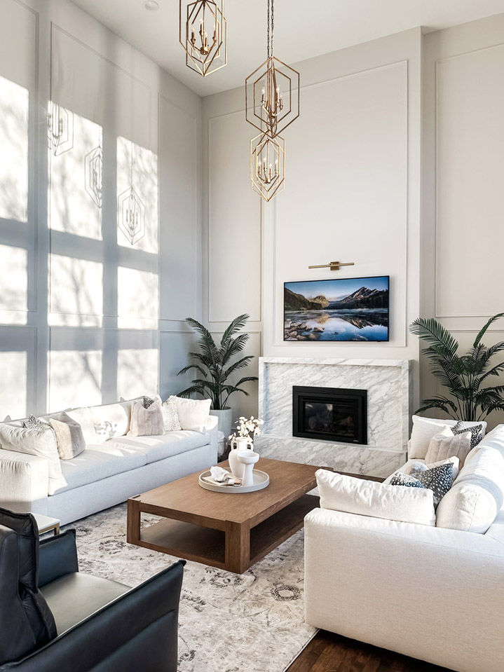 Stunning interior redesign of a living room by the Firelight Style House. The large white walls now have molding on them to create texture. The sun is coming in from the right and adds wonderful shadows of the new ceiling-mounted hanging lights that are bronze and diamond-shaped. There are two new, clean, and elegant white couches with white pillows. A newly designed marble fireplace was surrounded by a large landscape picture above the fireplace. Its interior was beautifully redesigned and imagined very well.