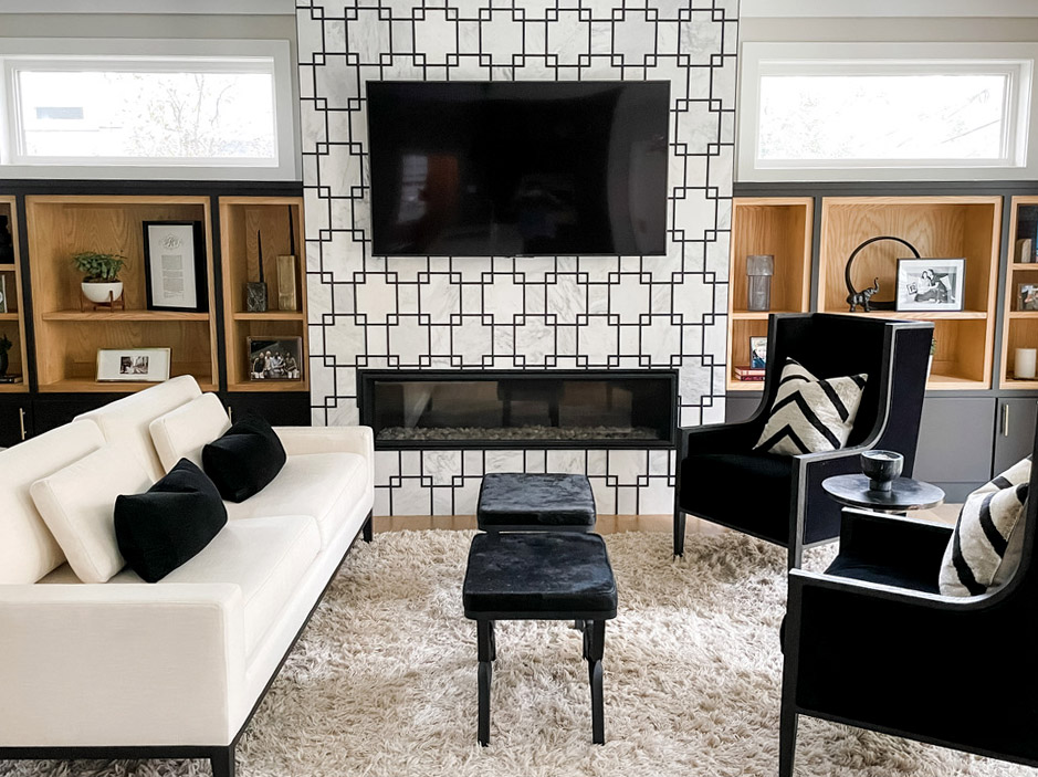 A black and white themed living room offers bold contrast and crisp styling. The focal point is a feature wall with a black geometric tile pattern framing a linear fireplace and wall-mounted TV. On either side, built-in open shelving in natural wood tones displays books, photos, and curated décor. A white modern sofa with black throw pillows pairs with two high-back black armchairs and a thick shag area rug for a striking and textural aesthetic.