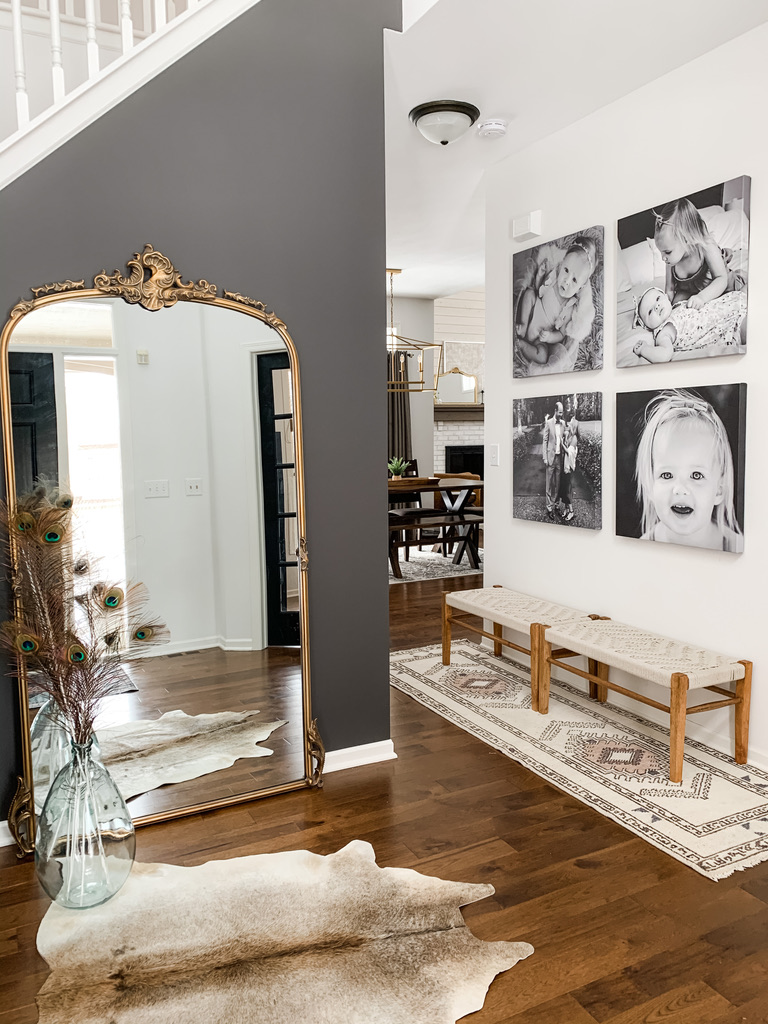 A transitional entry space showcases a dramatic dark accent wall with a full-length ornate gold mirror, reflecting natural light and a cowhide rug on warm hardwood floors. On the adjacent white wall, a series of black-and-white family portraits adds emotional warmth. A carved wood bench with a neutral patterned cushion sits over a long bohemian runner, completing the cozy, artful entryway.