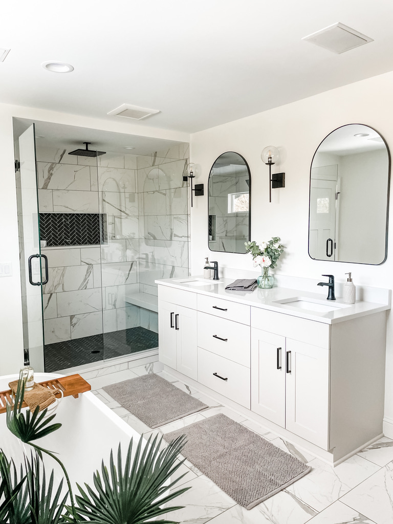 Bright and cohesive bathroom with a double vanity, arched mirrors, and a walk-in shower enclosed by frameless glass. Floor and wall tiles mimic the natural veining of marble, while greenery and soft, gray textiles add texture and comfort. This Minneapolis-St. Paul design reflects the interior design studio’s boutique approach to luxury living, blending function and style seamlessly.