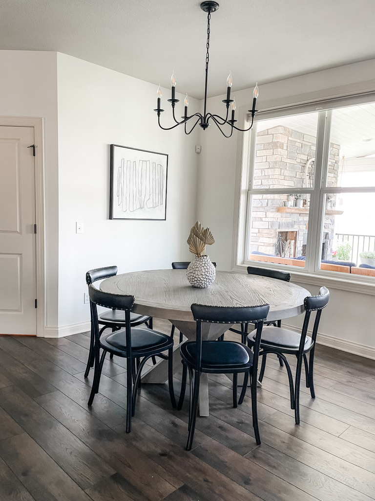 A transitional-style dining space features a round wood pedestal table with five navy upholstered chairs on dark hardwood floors. A matte black candelabra chandelier hangs overhead, and a neutral abstract line drawing adorns the wall. A large window reveals a view of an exterior stone fireplace. Designed by a boutique Minneapolis interior design firm, this space showcases how timeless elements can be elevated with custom touches and curated furnishings.
