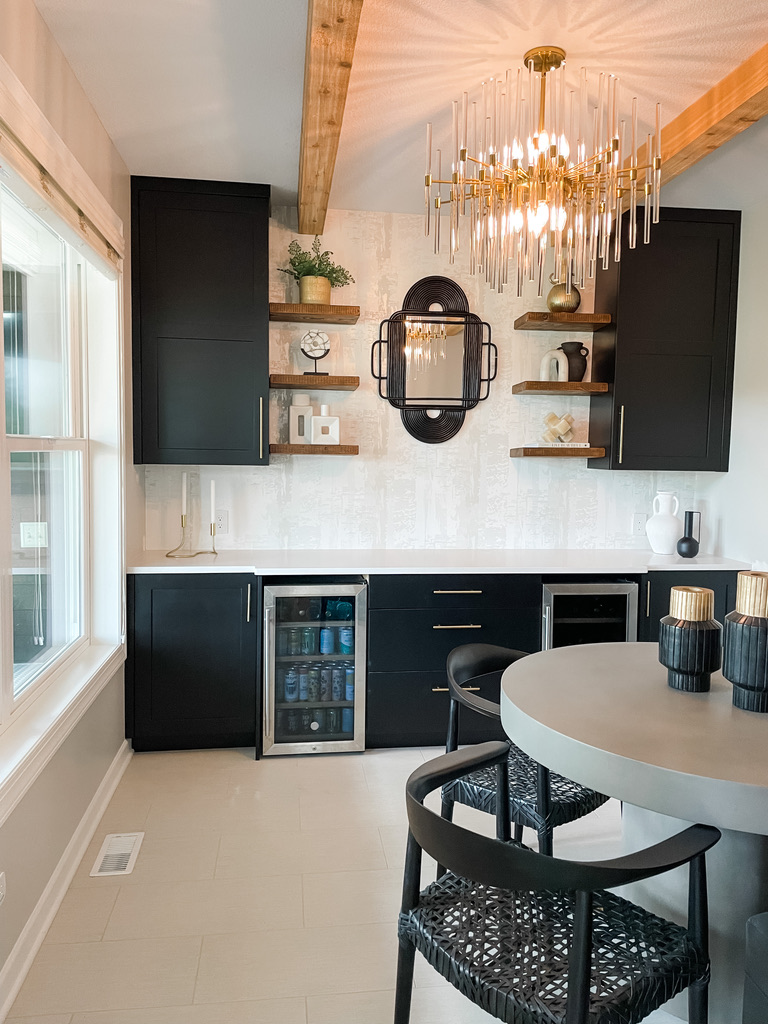 A chic built-in bar area features sleek black cabinetry, warm wood floating shelves, and a textured white wall. Dual beverage coolers are built into the lower cabinetry, while decorative objects, ceramics, and greenery accent the shelves. A black-framed mirror anchors the wall below a radiant gold-and-glass chandelier that reflects beautifully across the ceiling. The creative team at this Minneapolis interior design firm thoughtfully combines modern luxury with personalized styling, creating functional spaces that feel elevated yet livable.