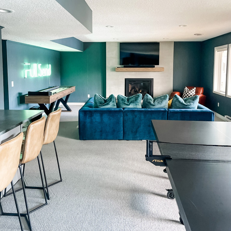 A photo of an interior redesign by the interior design team at Firelight Style House. The image looks directly into a room with a fireplace in the background wall, above it a TV mounted to the wall. The room has a soft blue-green color, a blue couch in the middle, a ping-pong table in teh foregound right and a small bar with three barstools on the left.