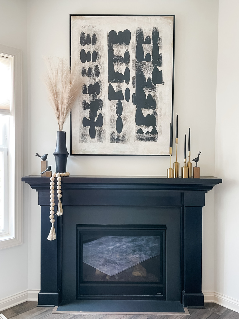 A dramatic black mantel anchors this modern fireplace vignette. Above it, a large abstract black and white painting adds a contemporary focal point. Decorative accents include a tall vase with pampas grass, gold and black taper candle holders, and sculptural bird figurines. A wood bead garland drapes casually over the edge of the mantel, softening the structured lines with organic texture.