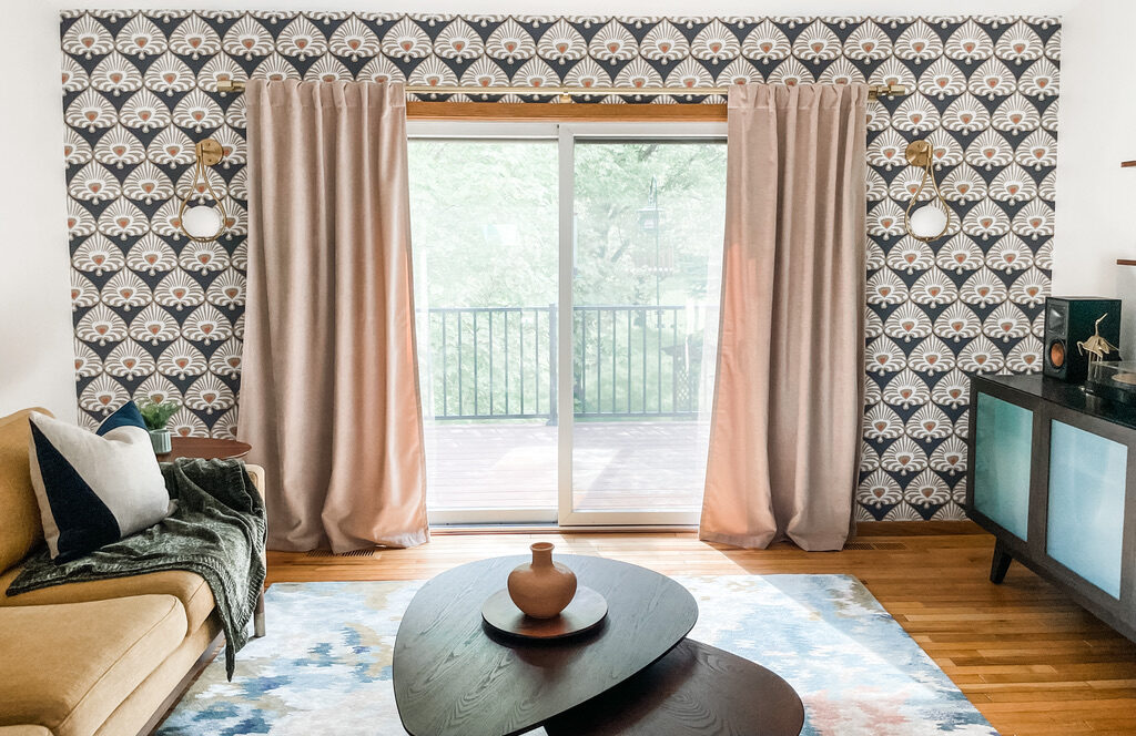 Eclectic and retro, this living room wall features dramatic geometric wallpaper in deep tones, paired with blush-colored curtains and a large sliding glass door that leads to a deck. The Art Deco theme continues with round wall sconces and a low, oval coffee table. A hint of personality shines through from the turntable and record cabinet to the right, infusing the space with a creative flair.