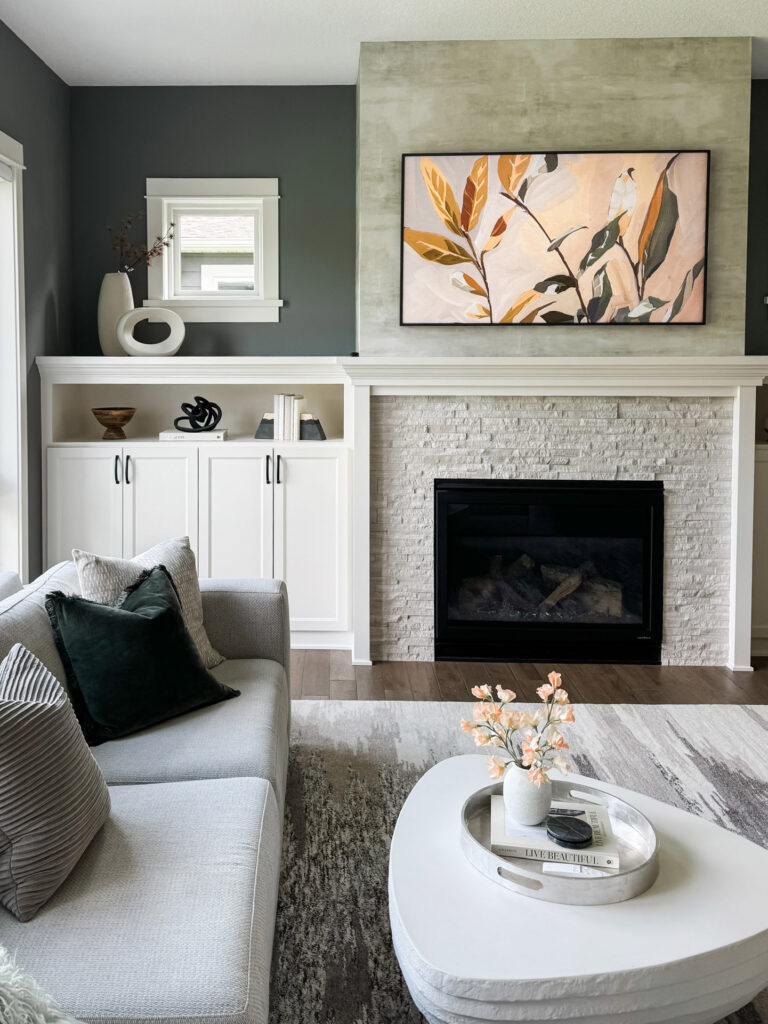 Modern luxury living room in a suburban Minneapolis home with deep green accent walls and a two-tone fireplace featuring white stone and a concrete finish. A gray sectional is styled with oversized velvet pillows, and a sculptural white coffee table with a floral arrangement completes the look. This space showcases the boutique interior design team’s expertise in blending soft textures with bold architecture to create a refined and livable aesthetic.
