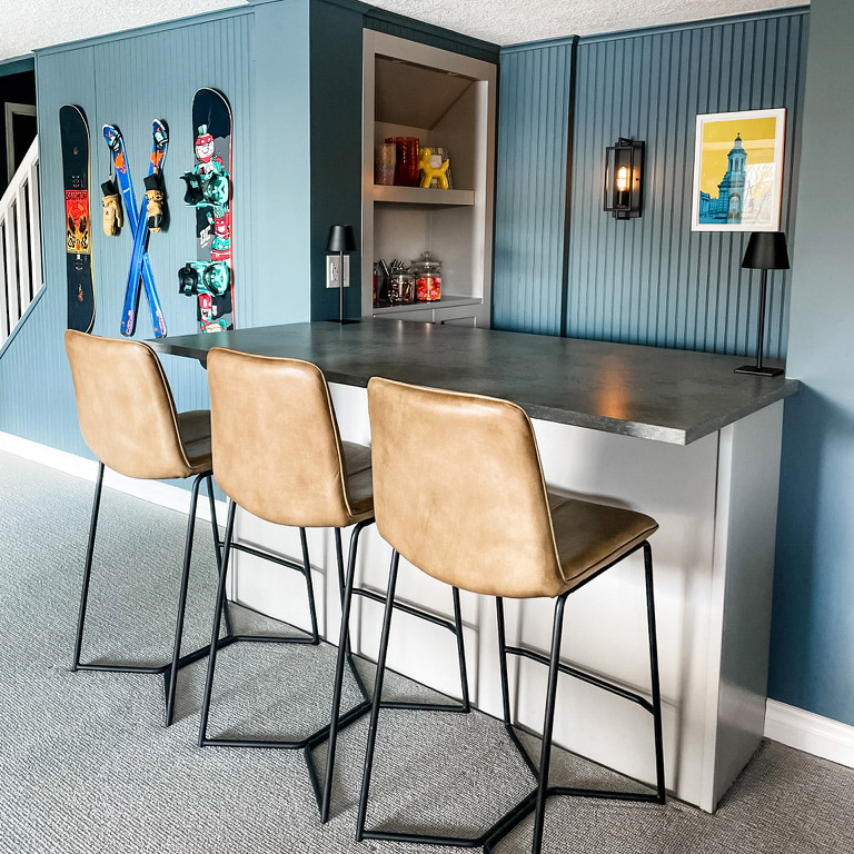Excellent interior design completed the bar. The walls are a light blue-green with ski equipment hanging on them. In the foreground, a small bar with three leather bar stools.