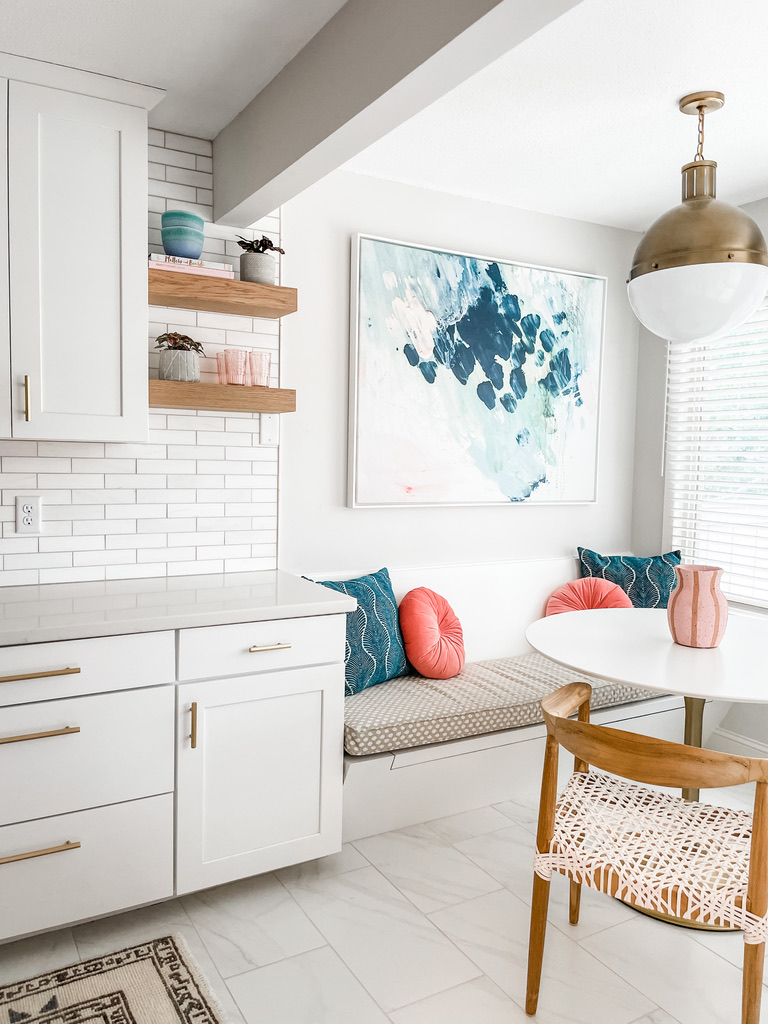 A modern eat-in kitchen nook with white cabinetry and gold pulls includes a built-in bench with light taupe polka-dot upholstery. The seating is accessorized with vibrant pillows in teal and coral. A round white tulip table and woven blush dining chair complete the space. A large abstract painting in oceanic tones brings an artistic touch, while a contemporary pendant light with a gold canopy and white glass shade hangs overhead. Natural light pours in from a nearby window with white blinds.