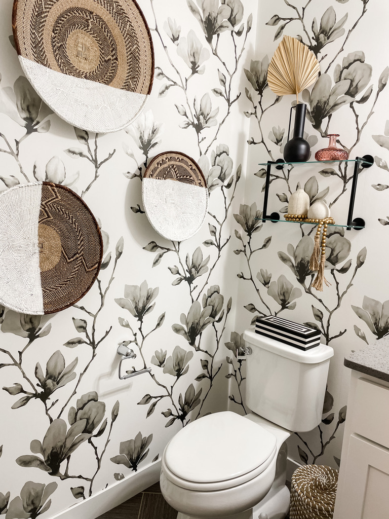 Eclectic powder room with hand-painted floral wallpaper in grayscale tones, paired with woven African baskets and modern decorative accents. A white toilet sits beneath floating glass shelves with vases, a palm fan, and beaded accessories. This Twin Cities design seamlessly combines global flair with local craftsmanship in a compact yet impactful space.