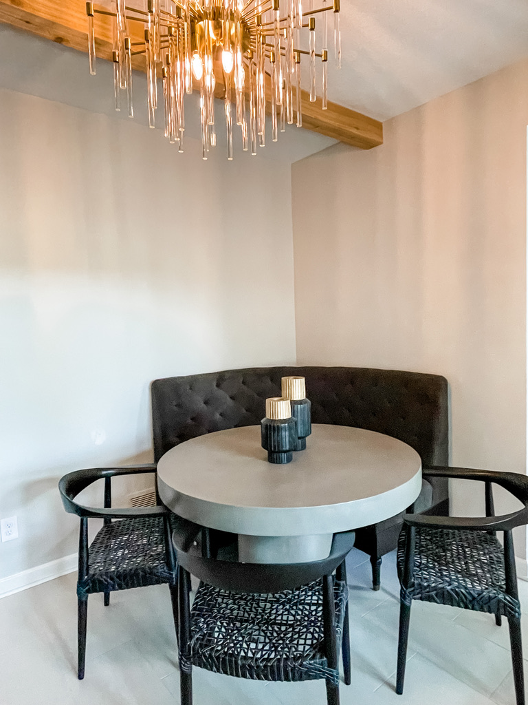 Cozy dining nook with a round gray table, black woven chairs, a curved banquette, and a sculptural glass chandelier beneath exposed wood ceiling beams.
