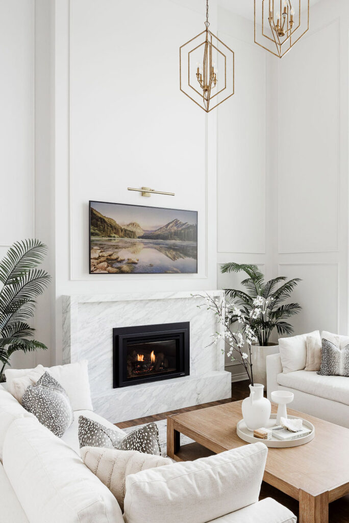 Stunning interior redesign of a living room by the Firelight Style House. The large white walls now feature molding to add texture. The upper middle of the pictures shows two hanging lights that are bronze and diamond shaped, very modern. There are two new, clean, and elegant white couches with white pillows. A large landscape picture above the fireplace surrounded a newly designed marble fireplace. Its interior was beautifully redesigned and imagined very well.