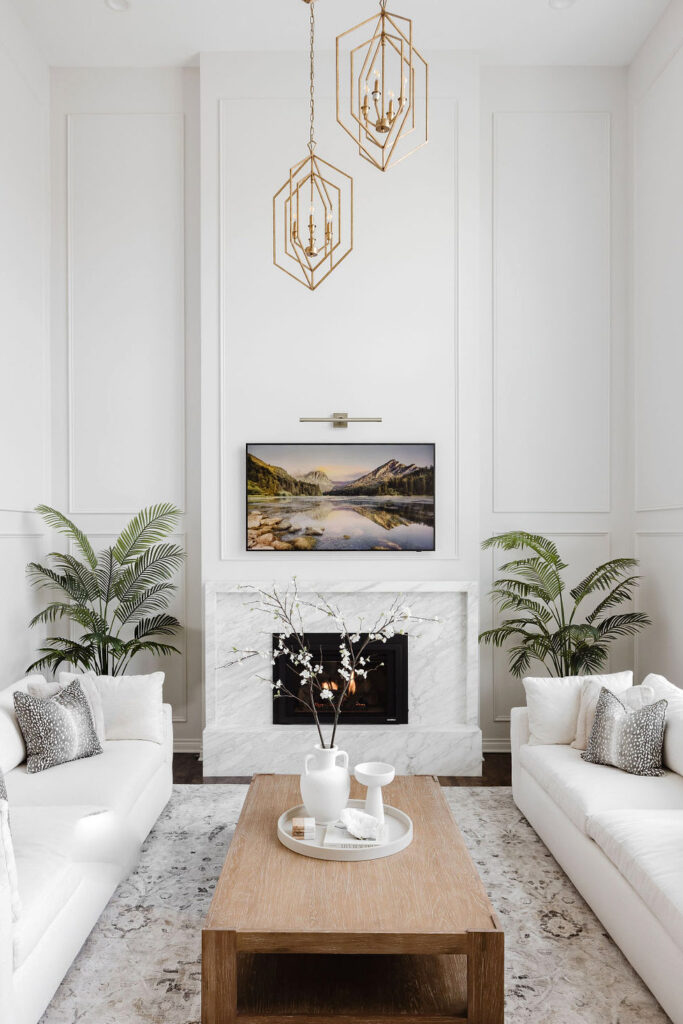 Stunning interior redesign of a living room by the Firelight Style House. The large white walls now feature molding to add texture. The upper middle of the pictures shows two hanging lights that are bronze and diamond shaped, very modern. There are two new, clean, and elegant white couches with white pillows. A large landscape picture above the fireplace surrounded a newly designed marble fireplace. Its interior was beautifully redesigned and imagined very well.