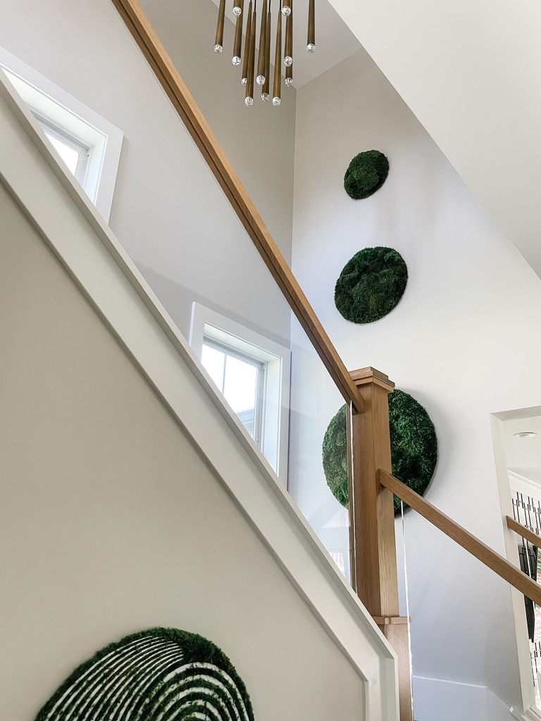 A close-up of a modern staircase in a Twin Cities home, showcasing natural wood railing and sculptural moss art mounted on the white wall. Above, a striking gold and crystal chandelier makes a bold artistic statement in the vertical space, a creative concept by the interior design team at Firelight Style House.