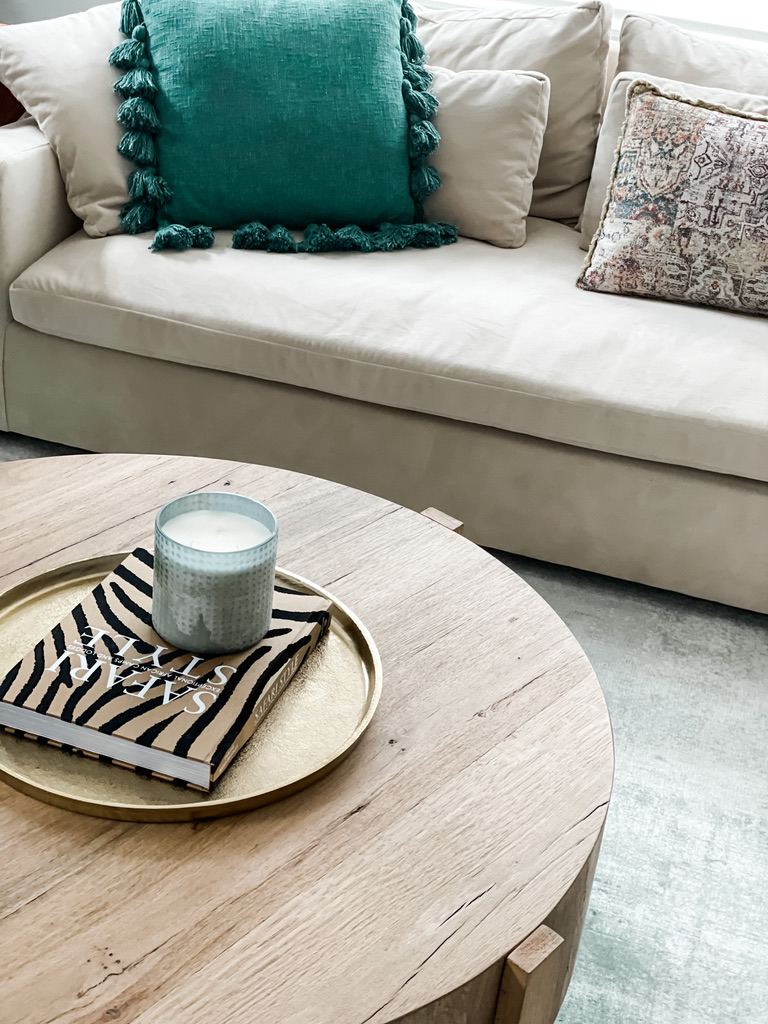 This close-up captures a cozy corner of a neutral-toned sofa accented by a large teal pillow with fringe tassels and a vintage-inspired patterned pillow. In the foreground, a round wood coffee table features a gold tray styled with a zebra-print book and a textured glass candle. Soft, layered textures and a muted palette create an inviting and lived-in living room feel.