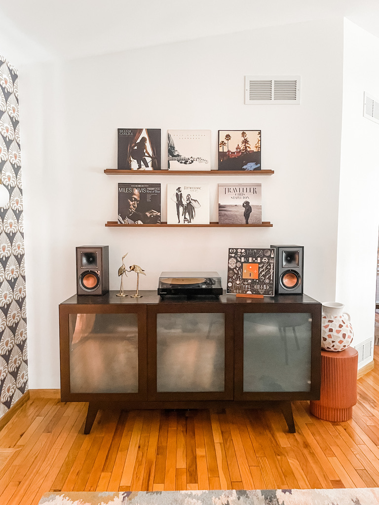 A mid-century modern inspired media setup showcases a dark wood console with frosted glass doors, topped by a turntable, golden crane figurines, and two compact speakers. Above, two floating shelves display a stylish vinyl record collection featuring Miles Davis and Chris Stapleton. Warm honey-toned hardwood floors and retro wallpaper off to the side enhance the space’s cozy, vintage vibe.