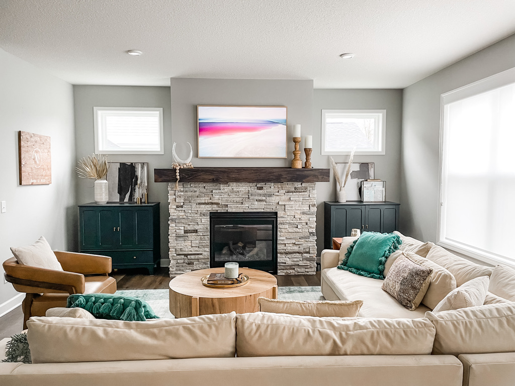 Cozy living room with neutral sectional seating, soft green throw pillows, and a circular natural wood coffee table. The fireplace is framed by a rustic stone surround, anchored by dark green cabinetry and soft natural lighting from clerestory windows. This inviting room is designed for comfort and style.