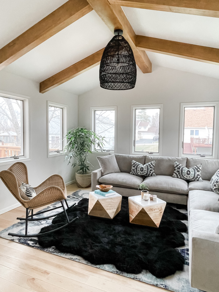 This view of a vaulted living room highlights the exposed natural wood beams and woven black pendant light. The neutral sectional is layered with tribal print pillows, flanked by a potted tree and a natural rattan rocking chair. The rich textures of a black shag rug and angular wooden coffee tables add grounded warmth to the sunlit space.