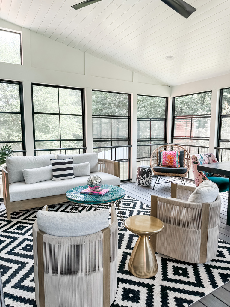 A wide view of a vibrant porch showcases the balance of texture and pattern. Two cream accent chairs with rope detailing sit opposite a modern white loveseat with black-and-white pillows. The diamond-patterned rug anchors the seating area, which also includes a teal-tiled table and a small gold side table.