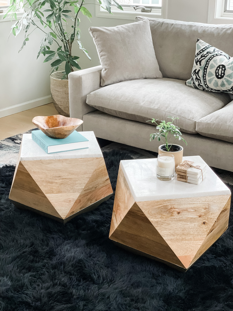 This close-up of a Minneapolis living room highlights two geometric wooden cube tables with faceted sides and white tops. The decor includes a wooden bowl, a plant, stacked books, and a candle, all placed atop a plush black area rug. The setup embodies a thoughtful balance between aesthetics and functionality, a skill at which the designers at Firelight Design Studio excel.