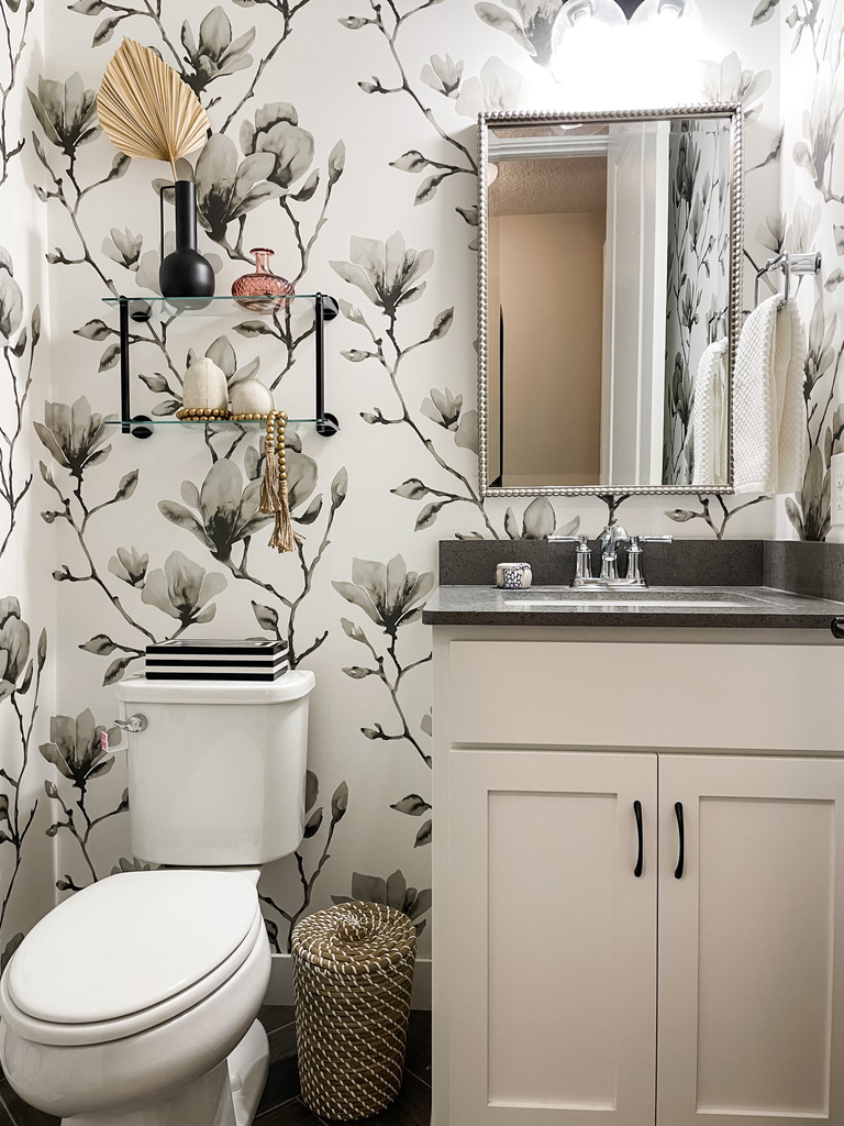 Charming powder room with gray stone countertop, white vanity, and a decorative silver-framed mirror. The floral-patterned wallpaper features soft grayscale blossoms, complemented by floating glass shelves with sculptural decor. Located in a Minneapolis-St. Paul area home, this design combines utility with artistic flair.