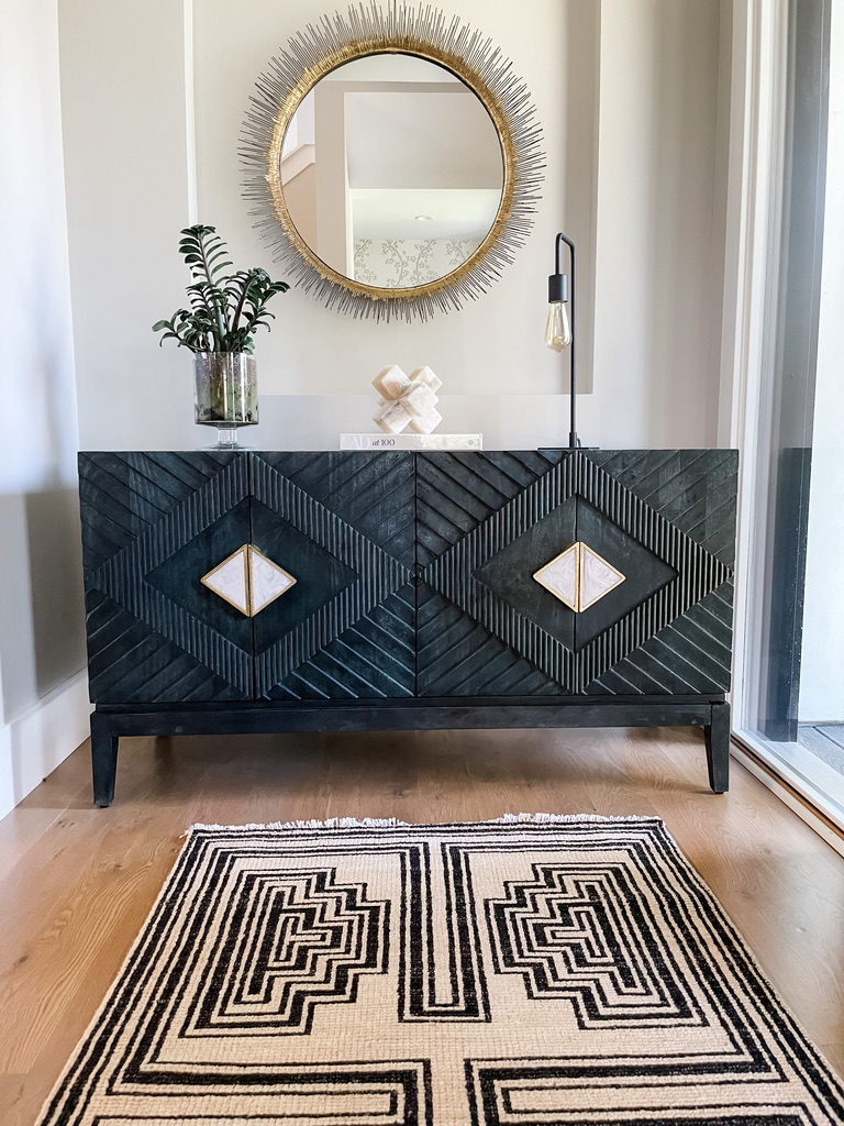 Entryway vignette in a home featuring a black wood console with gold and ivory geometric accents, topped with a round sunburst mirror and modern accessories. A black and cream patterned rug adds warmth to the wood flooring.