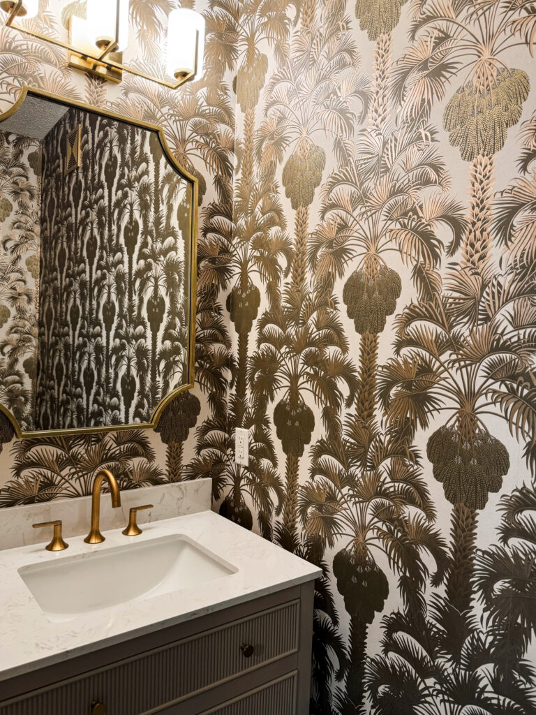 A bold powder room in the Minneapolis-St. Paul area features tropical-style wallpaper in metallic bronze palm fronds over a neutral backdrop, adding dramatic texture and pattern. The gold-framed mirror reflects the wallpaper’s symmetry, while a marble-look vanity top and brass faucet provide a luxurious finish.