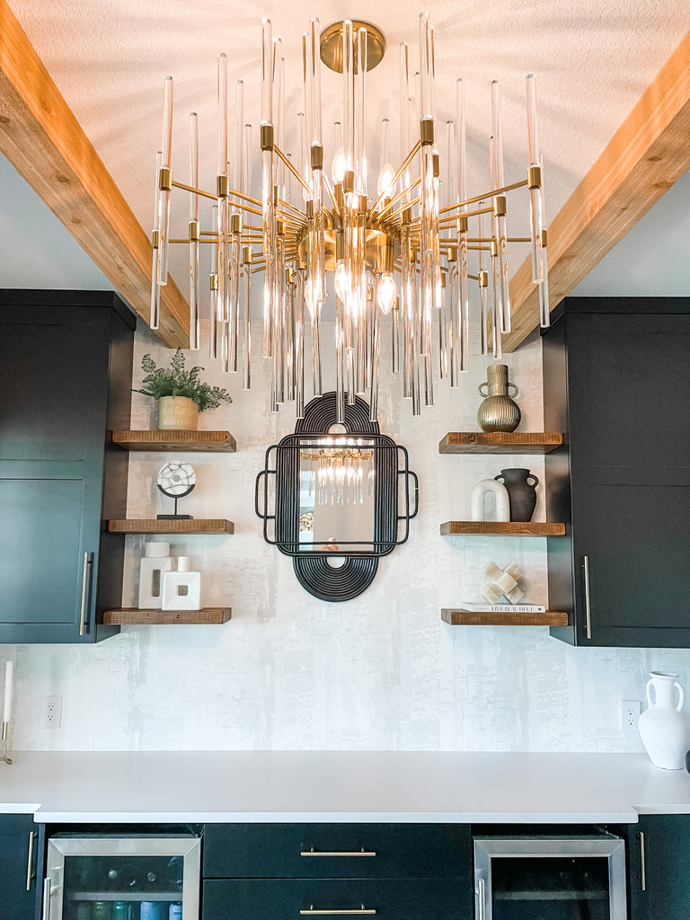 A striking modern chandelier made of gold rods and suspended glass tubes radiates from the ceiling, framed by exposed natural wood beams. Below, a custom-built beverage bar features matte black cabinets with gold handles and white quartz countertops. Floating wood shelves hold decorative vases, ceramics, and greenery, while a black metal-framed mirror serves as the central focal point on the textured white wall.