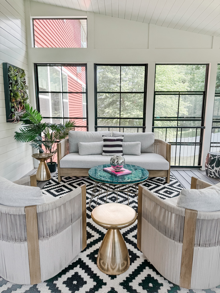 Conversation seating area in a Minneapolis-area three-season porch featuring a modern wood-framed loveseat and matching barrel chairs with wrapped cord accents, all arranged around a green tile coffee table and brass accent stools. This space showcases how timeless elements can be elevated with custom touches and curated furnishings that the Firelight Design Team creates.