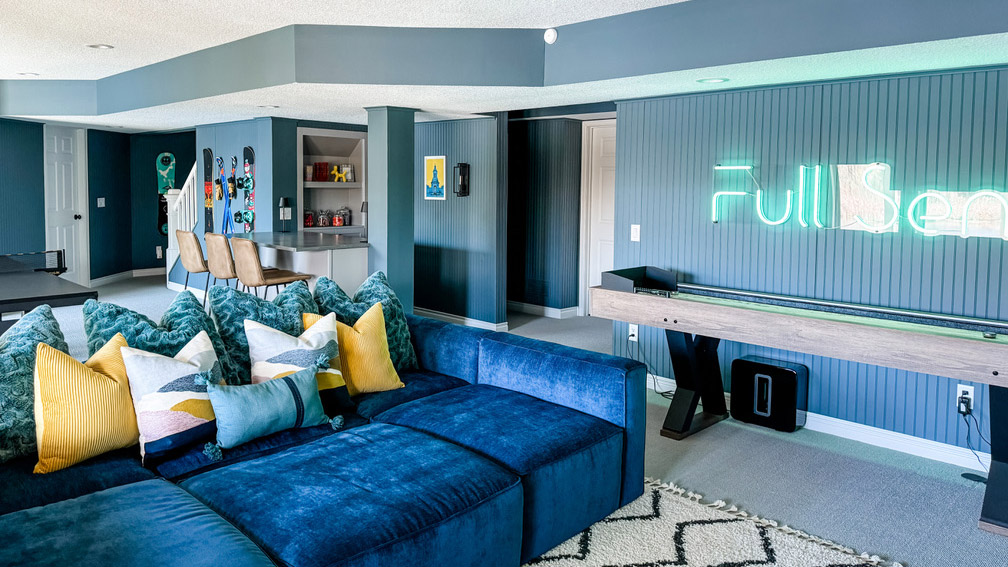 A lovely basement picture of a redesign by the Firelight Stylehouse interior design team. The walls are a soft blue-green, almost gray; there's a bright blue couch in the foreground with an array of pillows that are shades of blue, white, and yellow. In the background is a bar with three stools and a built-in bookshelf that's under the stairs. On the walls are downhill skis, three different snowboards, and a neon sign that reads "Full Send" in a fun neon green color.