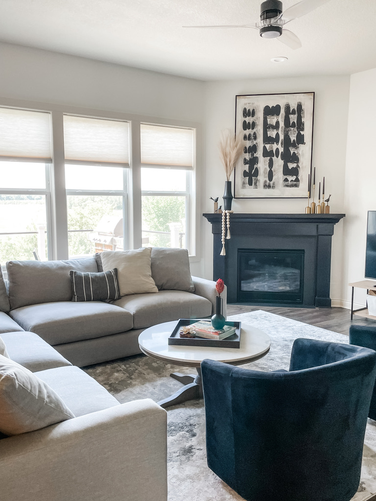 A sophisticated living space with a monochromatic palette in a Minneapolis suburb. A black-painted fireplace stands in stark contrast to the neutral walls and the natural light streaming through the large windows. A modern black-and-white abstract art piece hangs above the mantel. The room features a pale gray sectional sofa, a soft patterned rug, and a sleek circular coffee table topped with a sculptural green vase and dried botanicals. This living room reflects the boutique design team’s eye for blending modern form with inviting comfort, creating stylish, livable interiors across the Minneapolis metro.