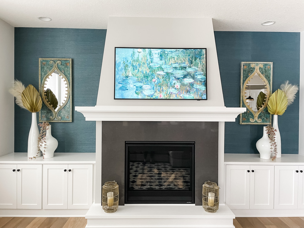 A modern living room fireplace in a Minneapolis-area home featuring a tall, tapered white chimney with a matte charcoal surround. Built-in white cabinetry flanks both sides of the fireplace, complemented by grasscloth-textured teal accent walls. Above each cabinet hangs an ornate mirror with an antique gold and green frame. Decorative white vases filled with dried palm leaves and pampas grass sit on either side, and a flat-screen TV displays Monet-style water lilies above the mantle.