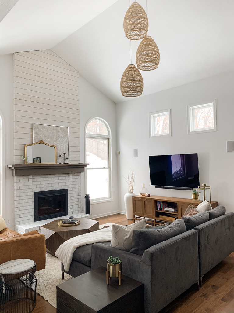 A cozy and light-filled living room in a Twin Cities home with a tall, vaulted ceiling. A white shiplap fireplace anchors the space, with a medium-gray brick surround and wood mantel. Natural woven pendants hang from the ceiling, enhancing the Scandinavian-inspired design. A charcoal sectional and a warm leather chair sit atop a soft, cream-colored rug. The large windows and warm wood flooring reflect a relaxed Minnesota lifestyle.