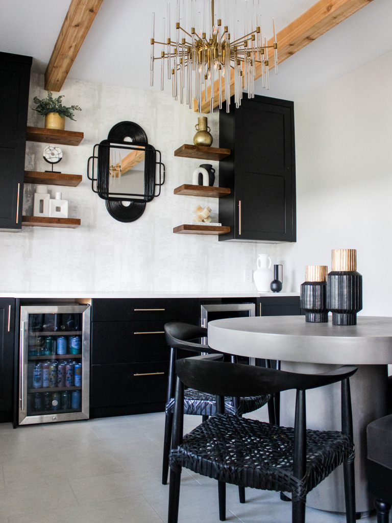 This stylish breakfast nook integrates matte black cabinetry with brushed gold hardware, a concrete pedestal dining table, and woven black dining chairs. Above, a sculptural brass chandelier with glass rods creates a dramatic visual anchor. The space features floating wood shelves with curated ceramics and greenery, a sculptural mirror, and a combination of natural wood beams and a soft, textured wall in white tones—capturing modern elegance in a Minneapolis interior.